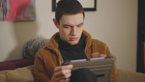 Young-Caucasian-Man-Sitting-On-Couch-Indoors-And-Using-Tablet---medium-shot