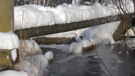 Person-is-crossing-snow-covered-log-bridge-over-icy-forest-river