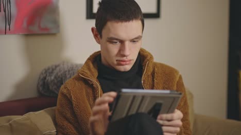 Young-Caucasian-Man-In-Brown-Furry-Jacket-Browsing-Internet-On-His-Tablet-While-Sitting-On-The-Sofa-In-The-Living-Room---Medium-Closeup-Shot