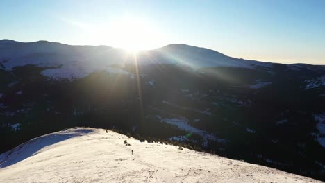 Luftaufnahme-Von-Wanderern,-Die-Mit-Schneeschuhen-In-Verschneiter-Berglandschaft-Auf-Wanderwegen-In-Felsigen-Bergen-Mit-Sonne-Als-Hintergrundbeleuchtung-Spazieren-Gehen