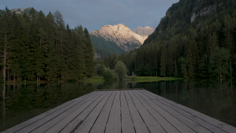 Alpine-mountain-lake-at-sunset,-Plansar-or-Plansarsko-jezero-in-Jezersko,-Slovenia,-wooden-pier-extending-above-the-water,-tilt-up-revealing-view