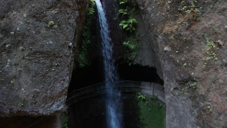 Female-runs-behind-waterfall-surrounded-by-rock-wall,-Levada-do-Moinho