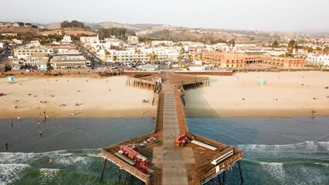 Aerial-forward-over-Pismo-wooden-pier-and-people-on-sandy-beach,-California