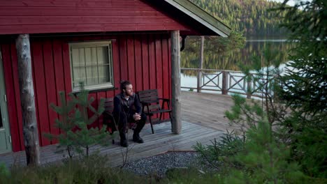 Man-Sitting-And-Looking-Around-At-The-Back-Of-Cabin-Near-Beautiful-Lake
