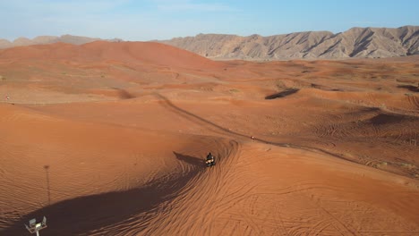 Drone-Camera-follows-a-tourist-riding-quad-bike-and-exploring-the-beauty-of-Sharjah-desert,-Meliha-mountains-in-the-background,-Desert-adventure-in-the-United-Arab-Emirates