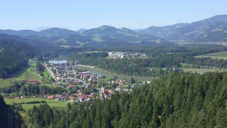 Aerial-approaching-charming-town-by-river-surrounded-by-forest-and-mountains
