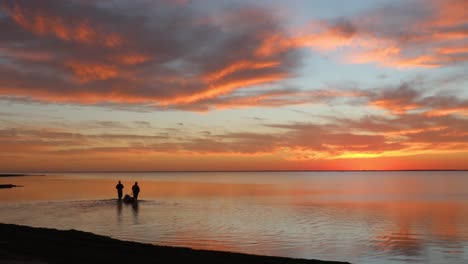 Zwei-Personen-Waten-In-Die-Ruhige-Mündung-Der-Laguna-Madres,-Während-Sie-Während-Eines-Farbenprächtigen-Sonnenuntergangs-In-Einem-Kleinen-Boot-Fanggeräte-Schleppen