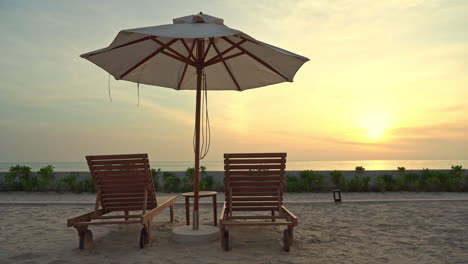 Nahaufnahme-Von-Zwei-Leeren-Liegestühlen-Und-Einem-Schattigen-Sonnenschirm-An-Einem-Sandstrand-Mit-Blick-Auf-Einen-Farbenfrohen-Sonnenuntergang-Am-Meer