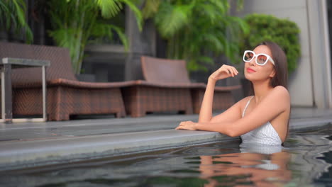Petite-Asian-Woman-in-Swimsuit-and-Sunglasses-Standing-in-Pool-Water-and-Looking-Around,-Full-Frame