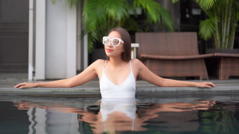 With-her-arms-stretched-out-along-the-edge-of-a-resort-pool,-a-pretty-young-woman-lounges-in-the-warm-pool-water