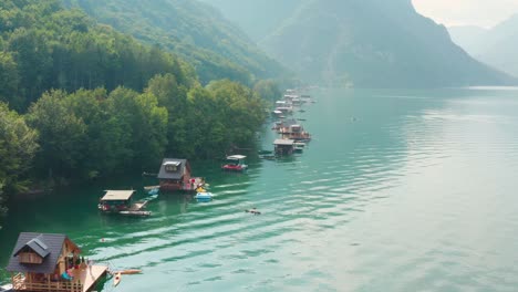 Aerial,-Wooden-houses-floating-on-Perucac-lake-surrounded-by-mountains-on-the-foggy-day