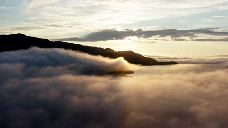 Drone-Flying-Above-The-Clouds-Over-The-Sea-With-Rocky-Mountain-In-Silhouette-On-A-Golden-Hour-Sunrise---wide-aerial-shot