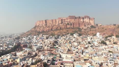 Blue-City-landscape-of-Jodhpur-surrounding-Mehrangarh-Fort-standing-above-cliff-in-Rajasthan,-India---Aerial-Pan-Reveal-shot
