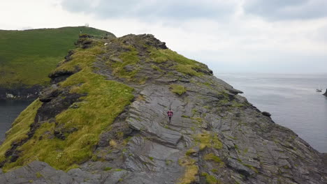 Hiker-climbing-and-ascending-rocky-coastal-headland-cliff,-Cornwall-UK,-aerial