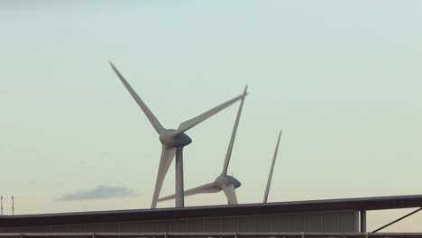 Windturbinen,-Die-Sich-über-Einem-Modernen-Außenfassadengebäude-Erheben,-Das-Strom-Erzeugt,-Wenn-Wolken-Bei-Sonnenuntergang-Vorbeiziehen