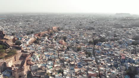 Jodhpur-blue-city-landscape-composed-by-small-buildings-surrounding-Mehrangarh-Fort-in-Rajasthan,-India---Aerial-wide-slide-shot