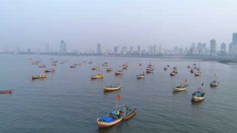 Volando-De-Lado-Con-Los-Barcos-De-Pescadores-De-Koliwada-Estacionados-En-Primer-Plano-Mientras-La-Ciudad-De-Mumbai-Se-Ve-En-La-Parte-De-Atrás-Bajo-Un-Smog-Como-Un-Clima-Brumoso
