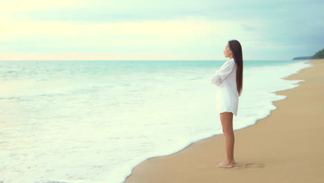 A-pretty-young-woman-stands-on-a-sandy-beach-looking-out-on-the-incoming-waves