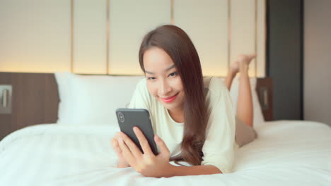 A-happy-young-woman-lying-on-her-stomach-on-the-bed-taps-a-message-on-her-cell-phone
