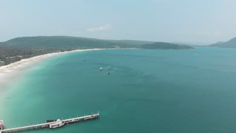 Wide-top-view-of-shoreline-landscape-encircling-paradisiac-bay-with-wooden-pier-in-Saracen-Bay-in-Koh-Rong-Sanloem,-Cambodia---Aerial-Wide-Panoramic-view