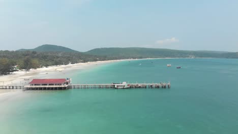 Old-Wooden-Pier-standing-above-calm-shallow-turquoise-water-dividing-the-enclosing-shoreline-in-Saracen-Bay-in-Koh-Rong-Sanloem,-Cambodia---Aerial-Fly-over-shot