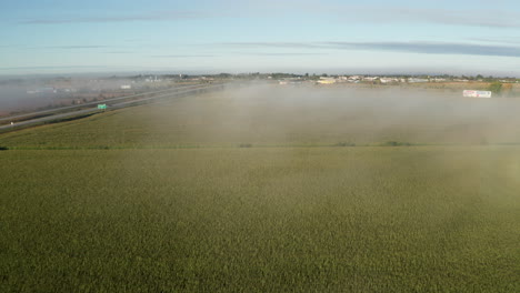 Light-covering-of-fog-hanging-over-farm-field-in-late-morning-seen-from-drone-flying-toward-town-on-horizon