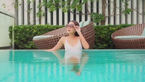 A-pretty,-young-woman-adjusts-her-sunglasses-as-she-leans-against-the-edge-of-the-pool