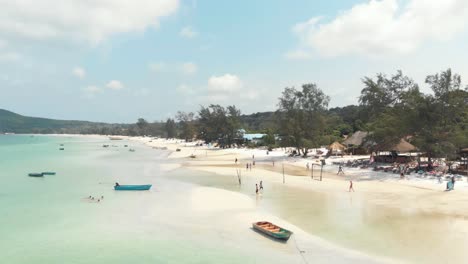 Populated-shallow-exotic-shore-with-tourists-and-fishing-boats-in-Saracen-Bay-in-Koh-Rong-Sanloem,-Cambodia---Aerial-low-angle-fly-over-shot