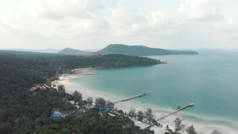 Wooden-beach-piers-entering-the-blue-shallow-sea-in-the-paradisiac-Saracen-Bay-in-Koh-Rong-Sanloem,-Cambodia---Aerial-wide-Fly-over-shot