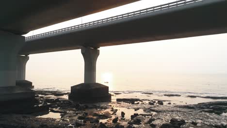 Fly-by-under-the-Bandra-Worli-Sea-Link,-revealing-a-fisherman-in-a-boat-with-the-sun-setting-in-the-back