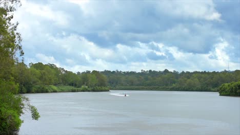 Jetskis-cruising-up-a-beautiful-and-picturesque-Australian-river,-with-its-heavily-wooded-banks,-under-cloudy,-heavy-skies