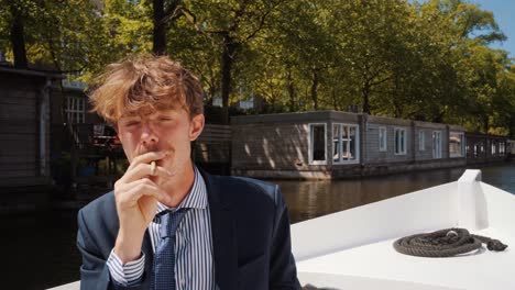 Male-student-with-mustache-smoking-weed-on-boat-in-Amsterdam-canals,-gives-joint