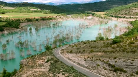 Car-moving-along-the-curved-road-next-to-Spain-mountain-cover-covered-with-water,-drone-covering-water-flow-along-Spain-ridges