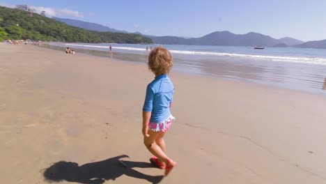 Junges-4-jähriges-Süßes-Mädchen,-Das-Frei-Am-Strand-Mit-Berghintergrund-Und-Ruhigem-Meer-Und-Klarem-Sand-Läuft