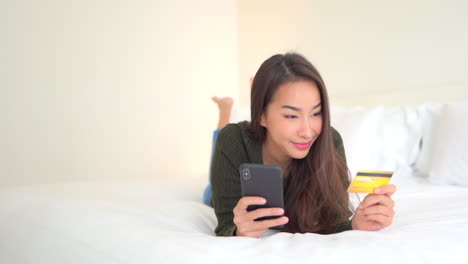 Junge-Asiatische-Frau,-Die-Auf-Bett-Liegt-Und-Kreditkarte-Und-Smartphone-Hält