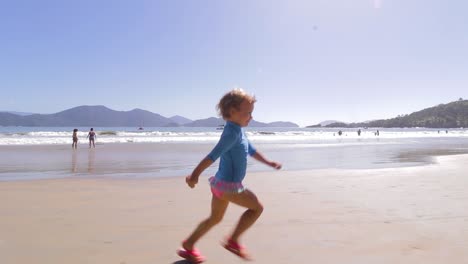 Kamera-Umkreist-Junges-Süßes-Mädchen,-Das-Frei-Am-Strand-Mit-Berghintergrund-Und-Ruhigem-Meer-Und-Klarem-Sand-Läuft