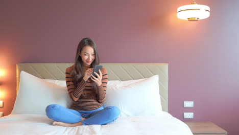 A-young-woman-sits-on-the-bed-in-her-hotel-suite-while-texting-on-her-mobile-phone