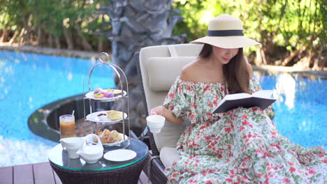 Elegant-woman-with-hat-and-floral-dress-sips-tea-while-reading-book-poolside