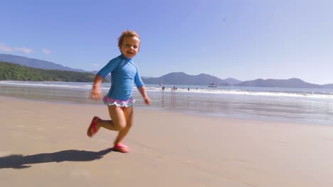 camera-orbiting-around-4-year-old-happy-young-cute-girl-running-freely-on-the-beach-with-mountains-background-and-calm-sea-and-clear-sand