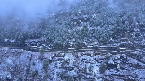 Aerial-footage-of-a-snowy,-scenic-byway,-winding-mountain-valley-road-during-a-snowstorm-with-pine-trees,-a-mountain-highway,-rocky-cliffs-and-forest-during-winter-on-a-cold,-blue-day