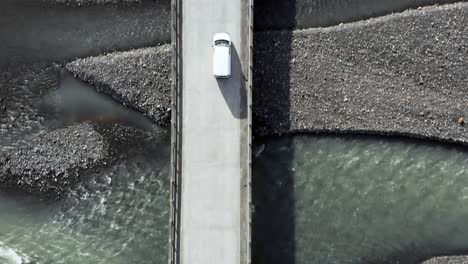 White-car-drives-across-bridge-over-meltwater-river-in-Iceland,-aerial