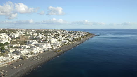 Drohne-Luftaufnahme,-Die-über-Schwarzen-Sandstrand-Auf-Der-Griechischen-Insel-Santorini-Fliegt,-Mit-Sonnenliegen-Und-Blauem-Meerwasser-In-4k