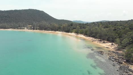 Rocky-golden-sandy-shoreline-with-lush-green-hills-encircling-the-shallow-turquoise-waters-of-M'pai-Bay,-Koh-Rong-Sanloem,-Cambodia---Aerial-low-angle-fly-over-shot