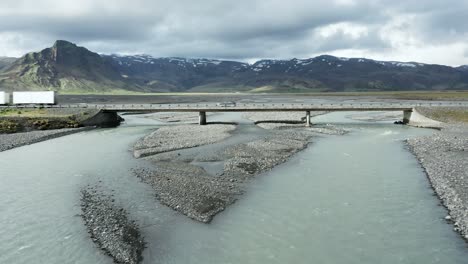 Aerial-flying-under-bridge-crossing-glacial-river-in-Iceland,-truck-on-road