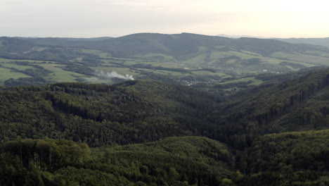 Aerial-of-forested-hills-and-smoke-rising-in-distance,-Central-Europe