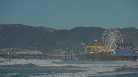 Mächtige-Wellen-Stürzen-In-Die-Küste-Und-Den-Pier-Mit-Dem-Berühmten-Riesenrad-Von-Santa-Monica-Und-Der-Landschaft-In-Der-Ferne---Zeitlupe