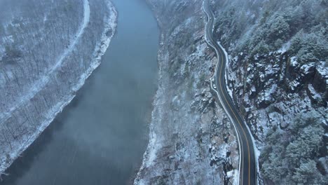 Aerial-footage-of-a-snowy,-scenic-byway,-winding-mountain-valley-road-during-a-snowstorm-with-pine-trees,-a-river,-mountain-highway,-rocky-cliffs,-and-forests-during-winter-on-a-cold,-blue-day