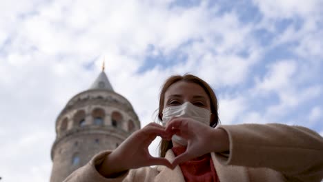 Beautiful-girl-wearing-protective-medical-mask-and-fashionable-clothes-does-heart-shape-with-hands-at-Galata-Tower-background