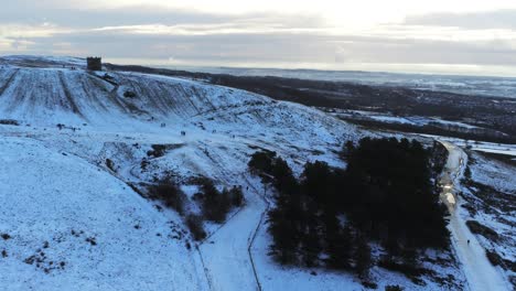 Snowy-Rivington-Pike-tower-Winter-hill-aerial-view-people-sledding-downhill-at-sunrise-slow-drift-left