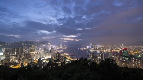 Wunderschöner-Zeitraffer-Der-Skyline-Von-Hongkong,-Victoria-Harbour-Bei-Sonnenuntergang-Mit-Hellen-Lichtern-Der-Stadt,-Schiffen-Und-Booten,-Die-Sich-Im-Hafen-Bewegen,-Und-Dicken-Wolken,-Die-Fließen